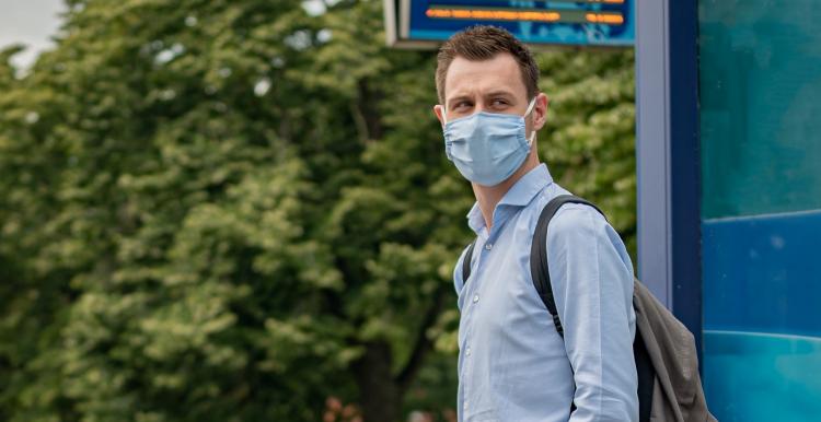 A man wearing a face covering and waiting at a bus stop