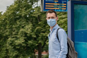 A man wearing a face covering and waiting at a bus stop